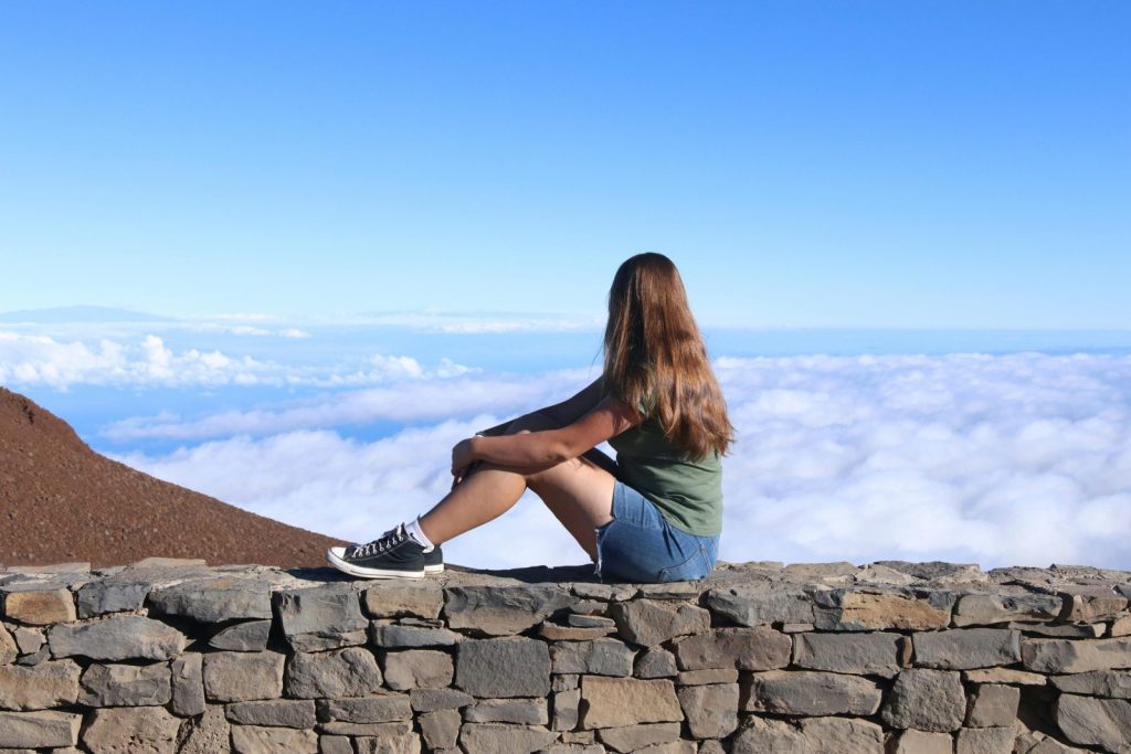 Young girl sitting on stone wall, enjoying scenic mountain view above clouds during day.
